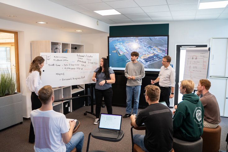 Drei Studierende präsentieren die Inhalte auf einem Whiteboard einer Gruppe von Kommilitonen.