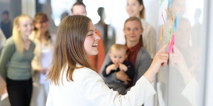 Gruppe Studierender löst Aufgabe am Whiteboard. Eine Studentin hält ein Baby im Arm.