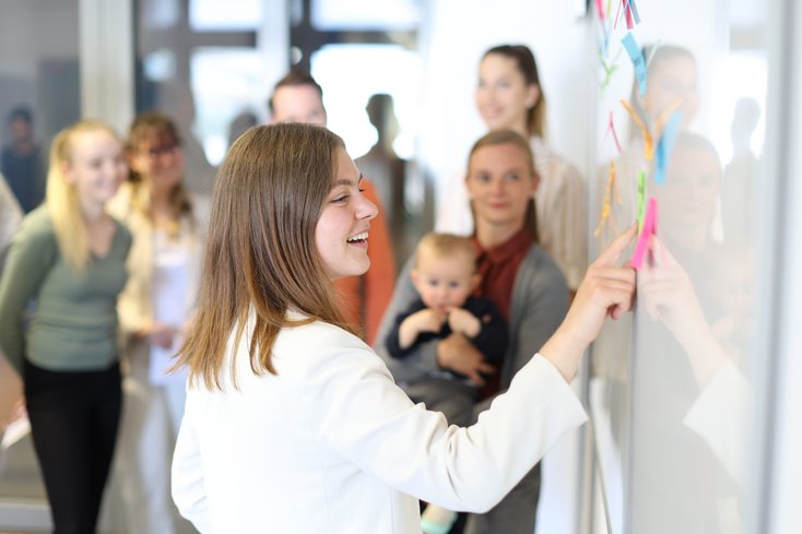 Gruppe Studierender löst Aufgabe am Whiteboard. Eine Studentin hält ein Baby im Arm.