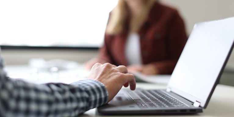 Eine Person tippt auf einem Laptop, der auf einem Tisch in einem hellen Raum steht. Im Hintergrund sitzt eine weitere unscharfe Person gegenüber, was auf ein Gesprächs- oder Arbeitsszenario hindeutet.