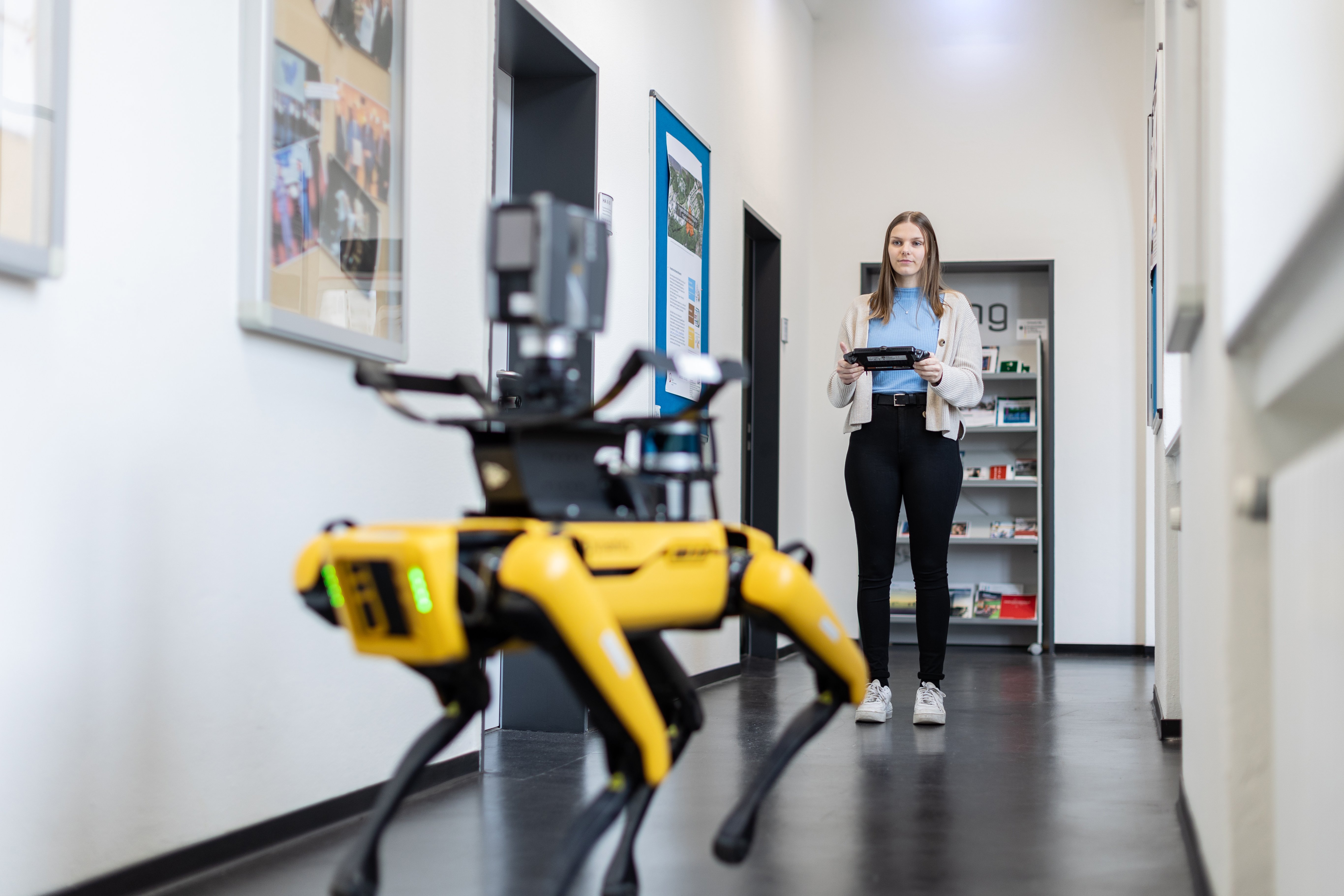 Eine Studentin steuert auf dem Flur einen Roboterhund mit einem Steuerungsmodul.