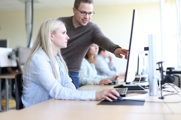 Stehender Mann erklärt sitzender Frau am Bildschirm etwas im Unterrichtsraum.