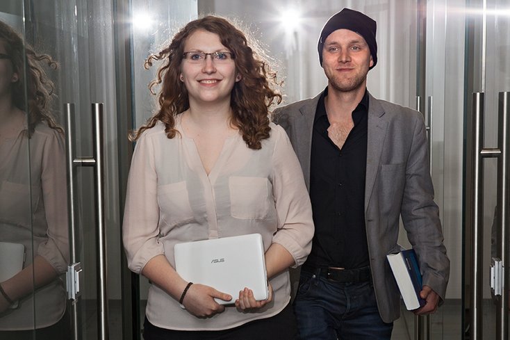 Junge Frau und junger Mann laufen nebeneinander einen Flur entlang auf den Betrachter zu.