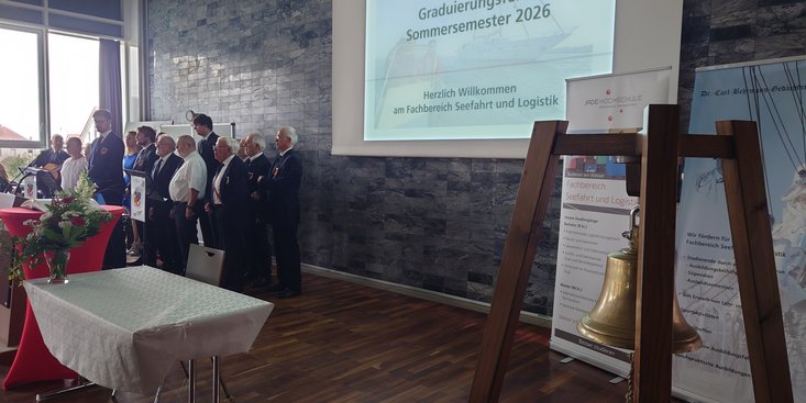 Im Vordergrund steht eine Schiffsglocke, im Hintergrund  ist der Shanty Chor und eine Präsentation zu sehen, auf der "Herzlich Willkommen" steht. 