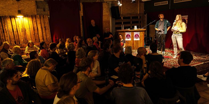 Zwei Personen stehen in einer Kneipe auf der Bühne. Vor ihnen sitzen viele Zuhörerinnen und Zuhörer.