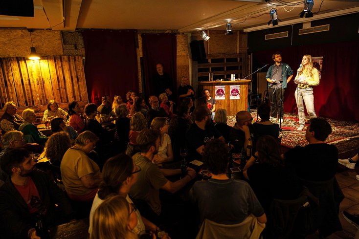 Zwei Personen stehen in einer Kneipe auf der Bühne. Vor ihnen sitzen viele Zuhörerinnen und Zuhörer.