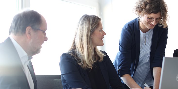 Eine Gruppe von vier Menschen in formeller Kleidung sitzt an einem Tisch um einen Laptop und arbeitet zusammen.