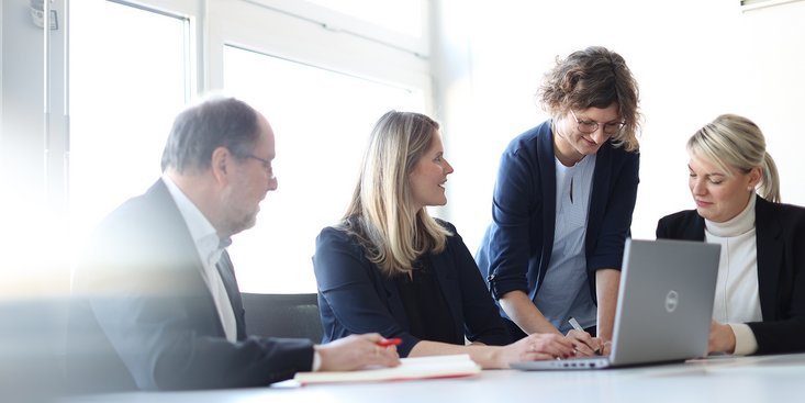 Eine Gruppe von vier Menschen in formeller Kleidung sitzt an einem Tisch um einen Laptop und arbeitet zusammen.