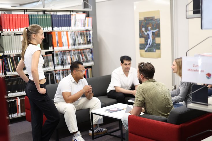 Vier junge Erwachsene sitzen in der Bibliothek in Oldenburg um einen Tisch und führen ein Gespräch, während im Hintergrund Bücherregale zu sehen sind. Eine Frau steht lächelnd daneben, während die anderen auf Sofas und Sesseln Platz genommen haben.