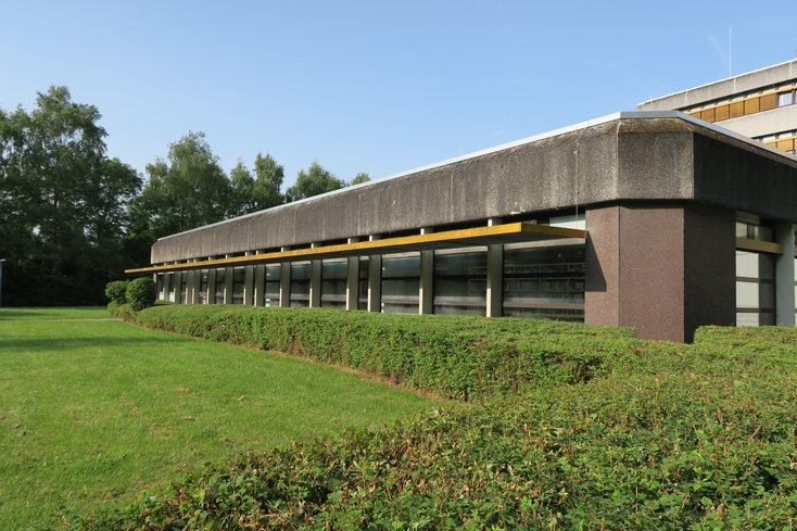 Das flache, funktionale Gebäude der Bibliothek am Campus Wilhelmshaven präsentiert sich mit einer dunklen Fassade, einer durchgehenden Fensterfront. Das Gebäude ist von einer gepflegten Rasenfläche und einer niedrigen Hecke umgeben, während im Hintergrund hohe Bäume zu sehen sind.