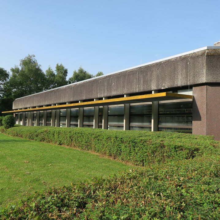 Das flache, funktionale Gebäude der Bibliothek am Campus Wilhelmshaven präsentiert sich mit einer dunklen Fassade, einer durchgehenden Fensterfront. Das Gebäude ist von einer gepflegten Rasenfläche und einer niedrigen Hecke umgeben, während im Hintergrund hohe Bäume zu sehen sind.