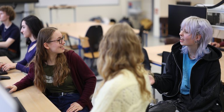 Drei junge Personen sitzen in einem Klassenraum an Computertischen und unterhalten sich, während sie sich einander zuwenden. Im Hintergrund sind weitere Schüler und Computerarbeitsplätze unscharf zu sehen.