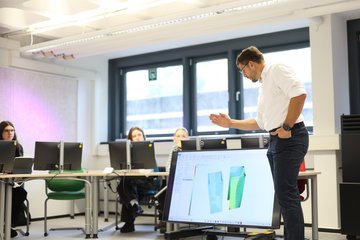 Dozent erklärt etwas am großen Bildschirm.