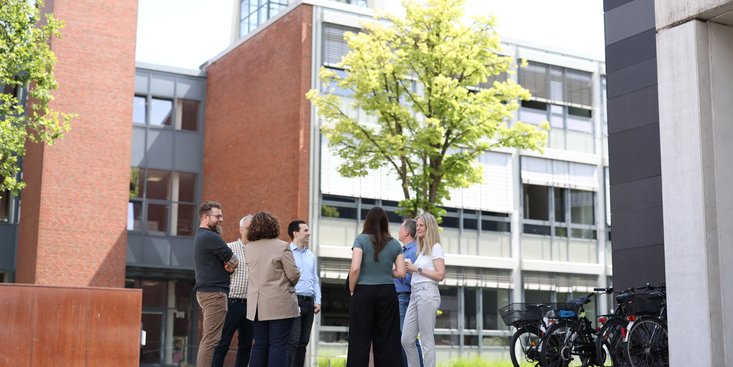 Menschengruppe unterhält sich stehend und gut gelaunt vor dem Hochschulgebäude.