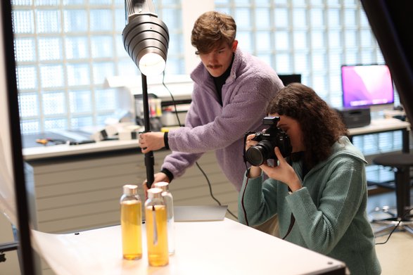 Zwei Studierende mit Fotokamera und Scheinwerfer im Fotostudio beim Fotografieren von Glasbehältern mit einer gelben Flüssigkeit