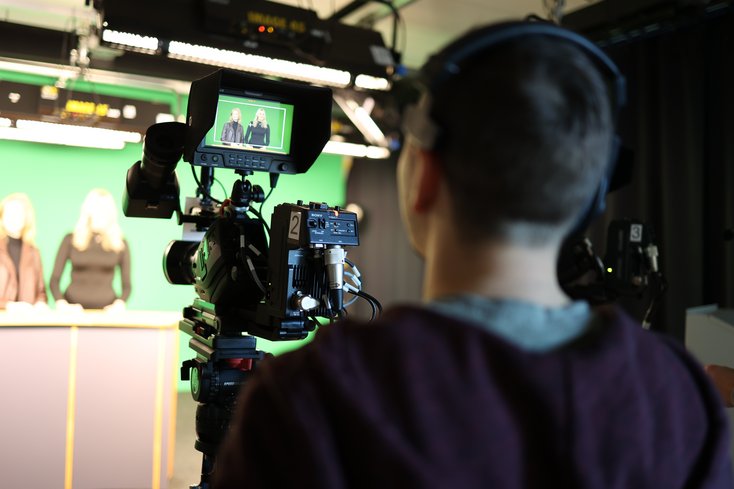 Studentin mit Filmkamera, Ansicht von hinten, im Hintergrund Lichttechnik und Moderationsbühne mit Greenscreen
