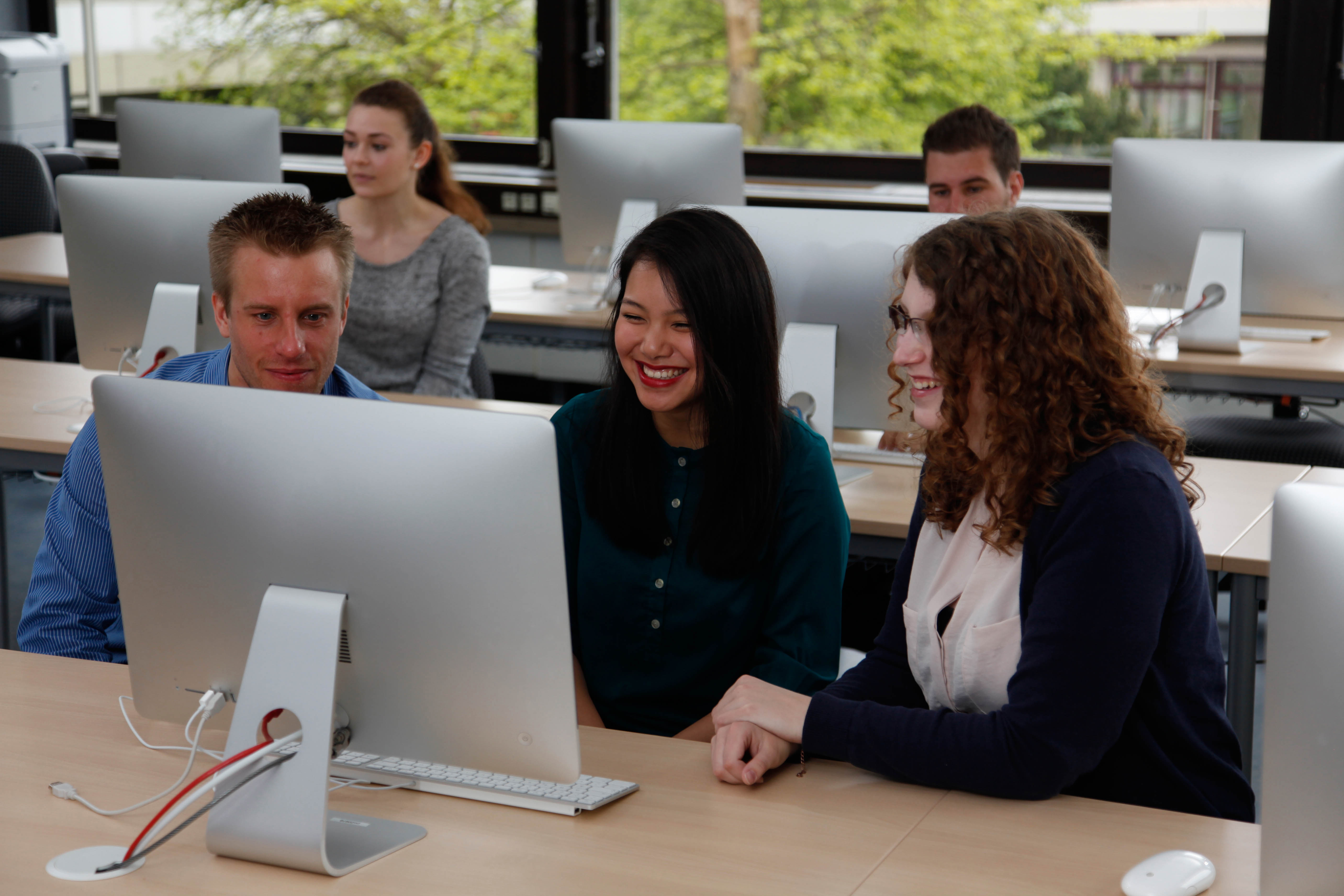 Im Vordergrund ein männlicher und zwei weibliche Studierende, die auf einen Bildschirm schauen. Im Hintergrund weitere Bildschirme und Studierende.