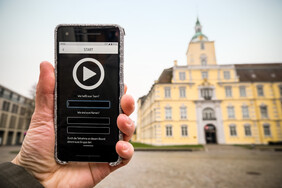 Eine Hand hält ein Smartphone, die App läuft und im Hintergrund ist das Oldenburger Schloss zu sehen.