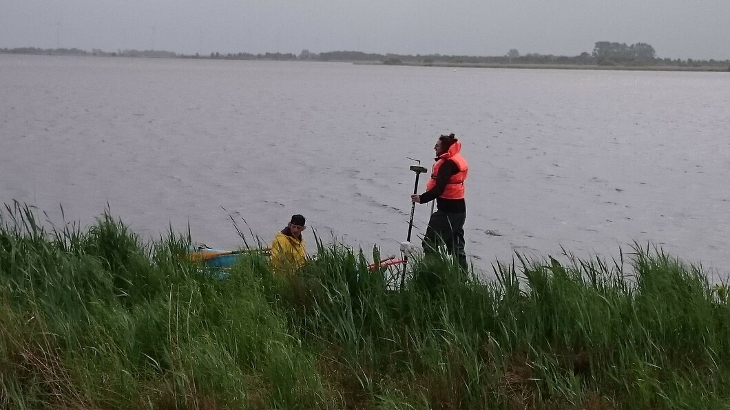 Zwei Studierende befinden sich bei regnerischem Wetter im Uferbereich eines Sees, eine GPS-Antenne wird festgehalten