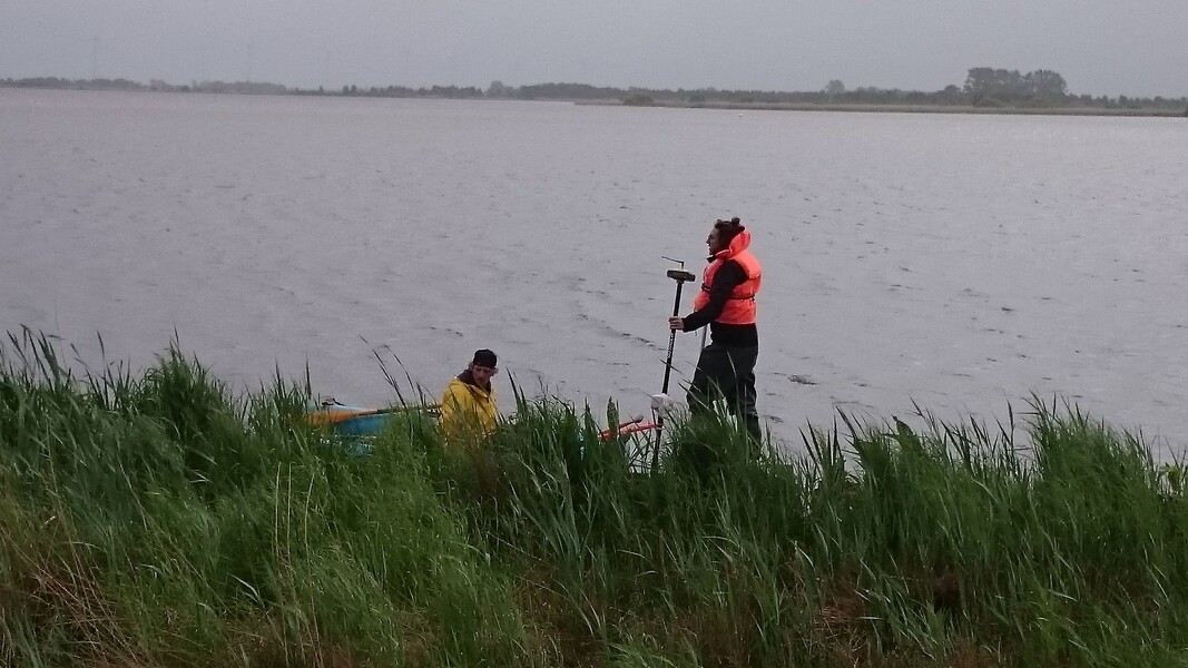 Zwei Studierende befinden sich bei regnerischem Wetter im Uferbereich eines Sees, eine GPS-Antenne wird festgehalten