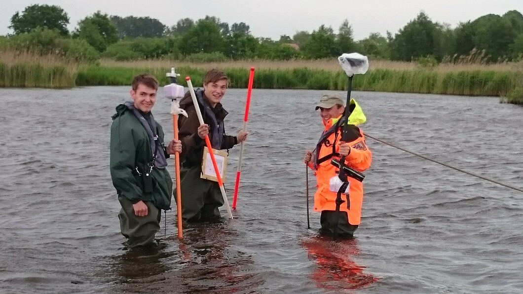 drei männlich gelesene Studierende stehen mit Wathosen im Wasser und haben Vermessungszubehör in der Hand