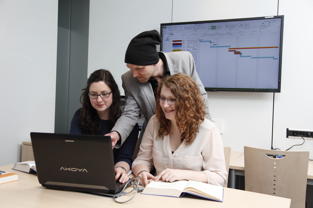 Drei Studierende sitzen über Laptop gebeugt und besprechen sich