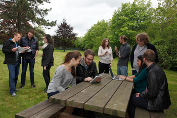 Gruppenarbeit im Freien, einige Studierende sitzen an einer Holzbank, einige andere stehen weiter entfernt