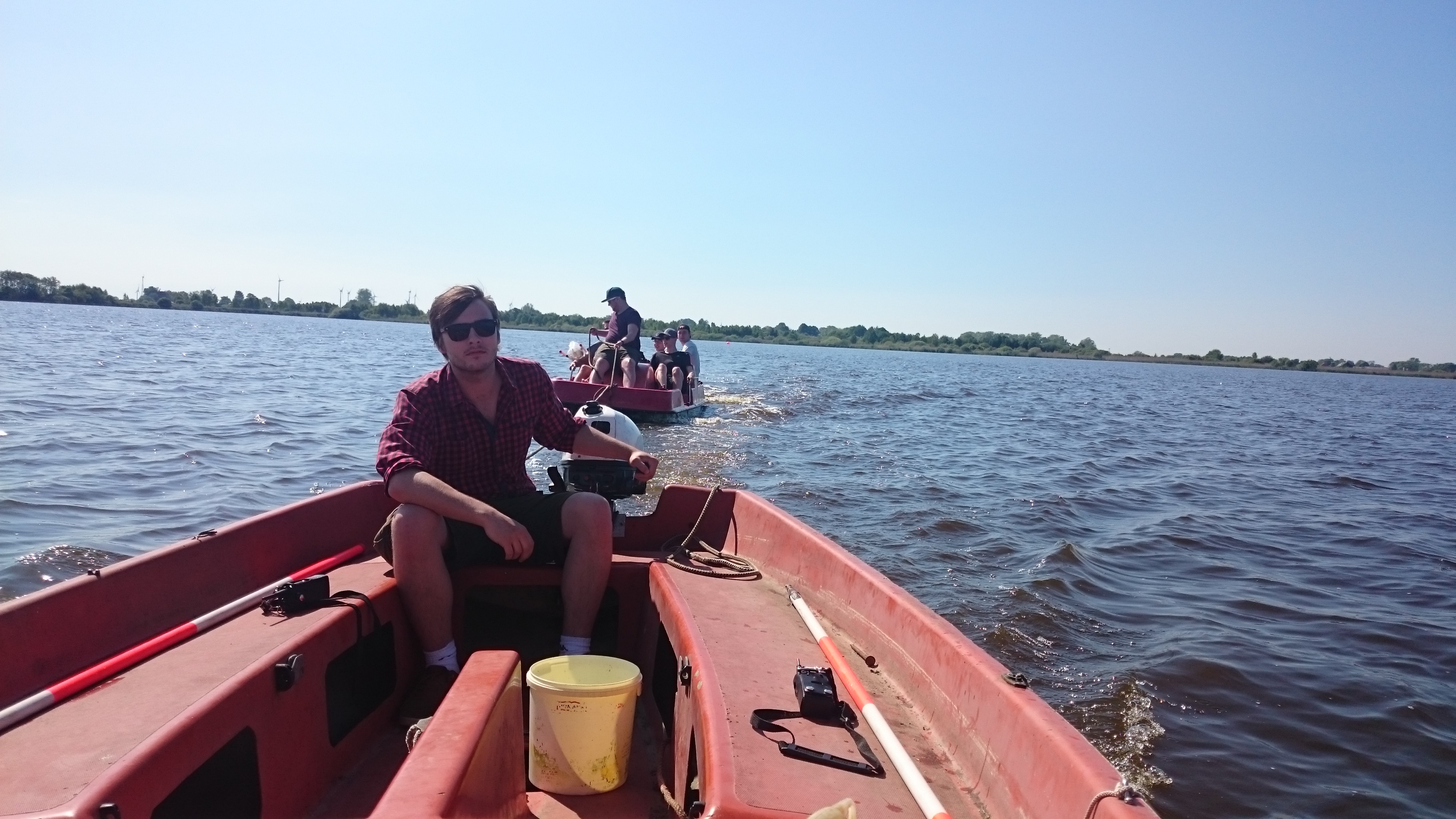 Im Vordergrund ist ein rotes Boot mit Heckmotor zu sehen, das eine Person steuert. Im Hintergrund ist ein kleines Boot mit mehreren Studierenden zu sehen, das vom Motorboot gezogen wird.